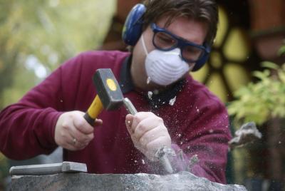 Mann im dunkelrotem Pulli, mit Mundschutz, Augen- und Gehörschutz, der mit Hammer und Meißel einen Stein bearbeitet. Mann im dunkelrotem Pulli, mit Mundschutz, Augen- und Gehörschutz, der mit Hammer und Meißel einen Stein bearbeitet.